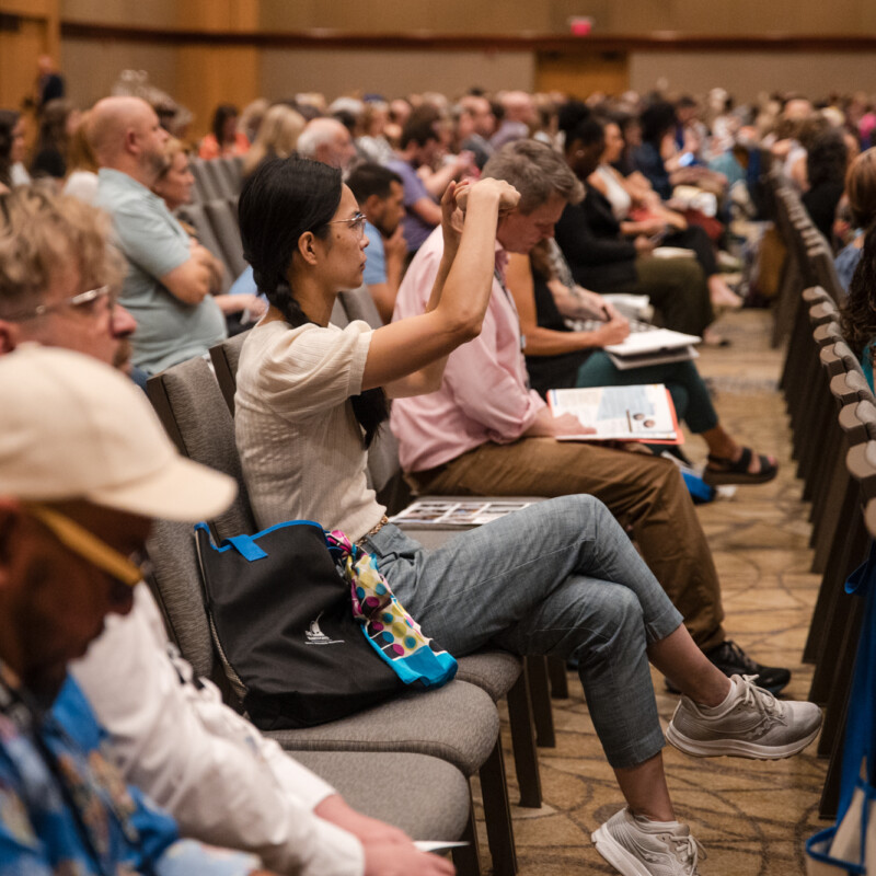 People attending a large session take notes while listening to a presenter (out of frame).