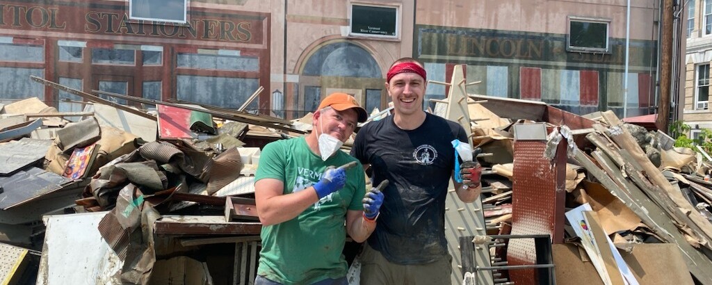 Two men pose in front of a large pile of flood debris