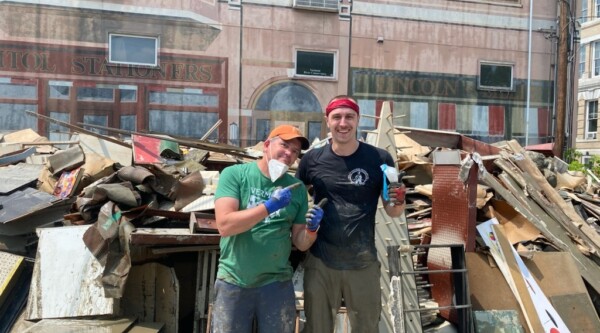 Two men pose in front of a large pile of flood debris