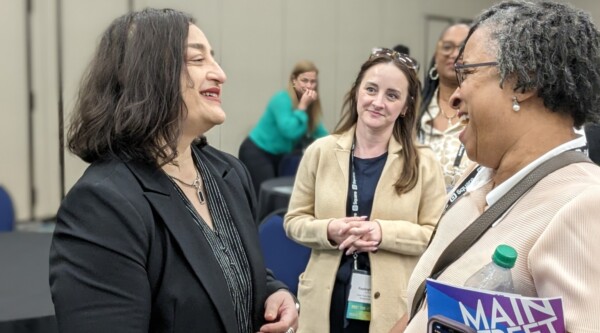 Two women shake hands and chat in a room filled with people networking.