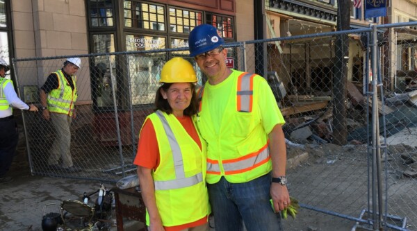 Two people wearing reflective vests and hard hats pose for a photo outside a historic building