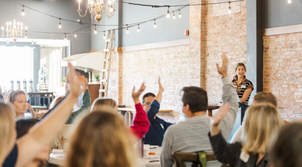 People seating around tables raise their hands; in the background a speaker holds a microphone.