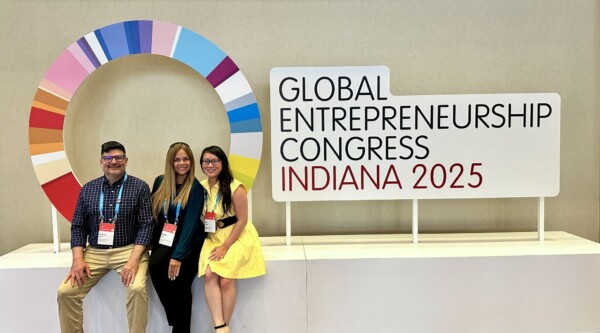 Un hombre y dos mujeres posan delante de un cartel con el logotipo del congreso mundial sobre iniciativa empresarial 2025.