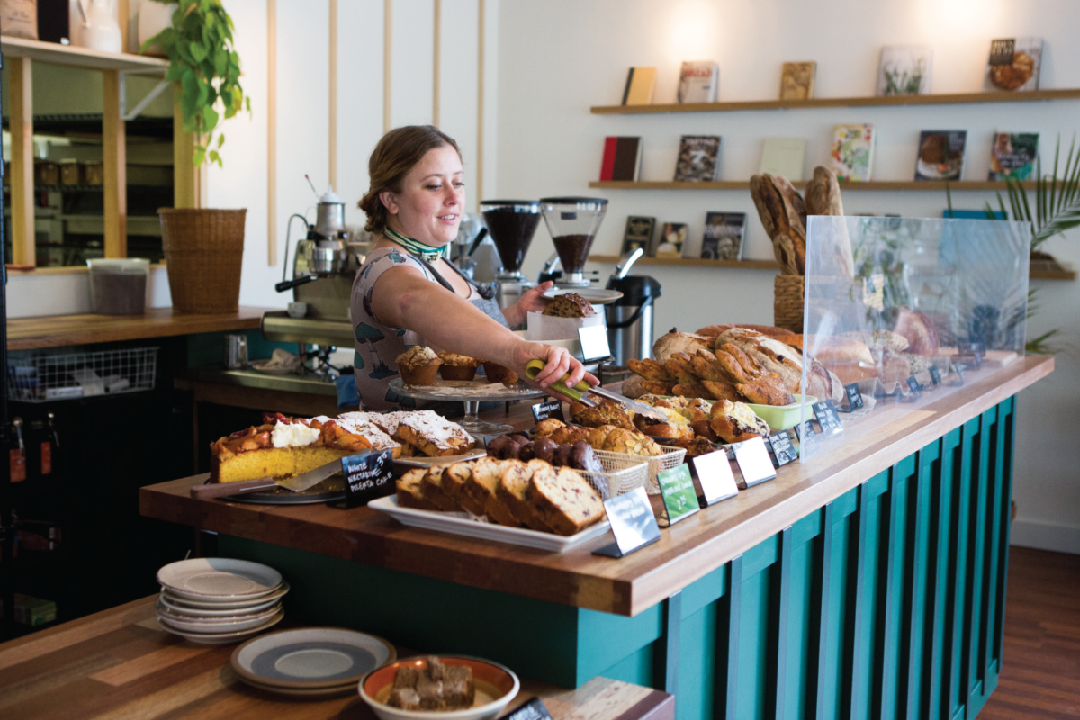 A woman behind a bakery counter uses tongs to pick up bread