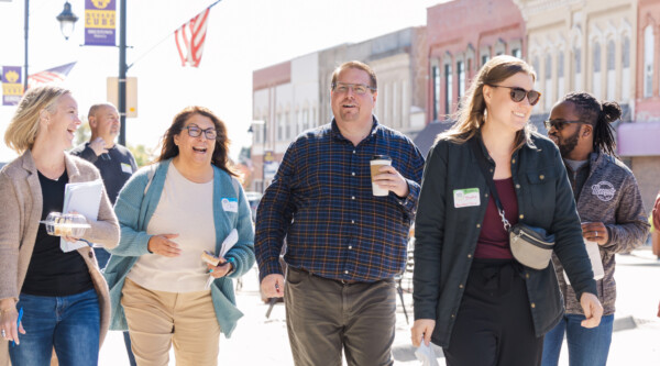 A group of adults laugh while walking through a downtown area with brick buildings and banners visible in the background.