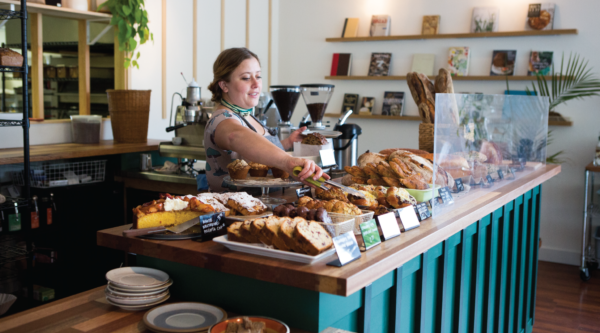 A woman behind a bakery counter uses tongs to pick up bread