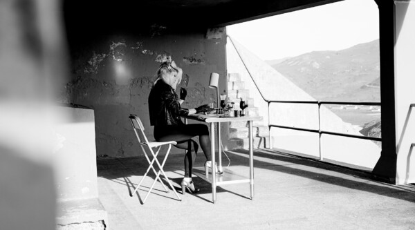 Black and white photo of woman wearing gloves and sitting at desk with perfume bottles.