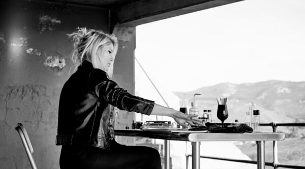 Black and white photo of woman wearing leather jacket sitting at desk with glass bottles.
