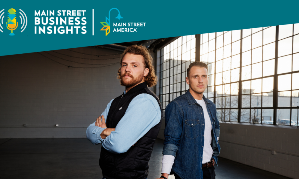 Two young men stand back to back in front of large industrial window.