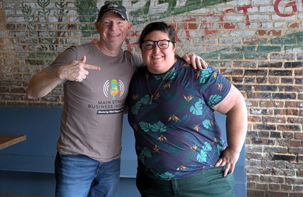 Man and woman smile with arms around one another in front of brickwall