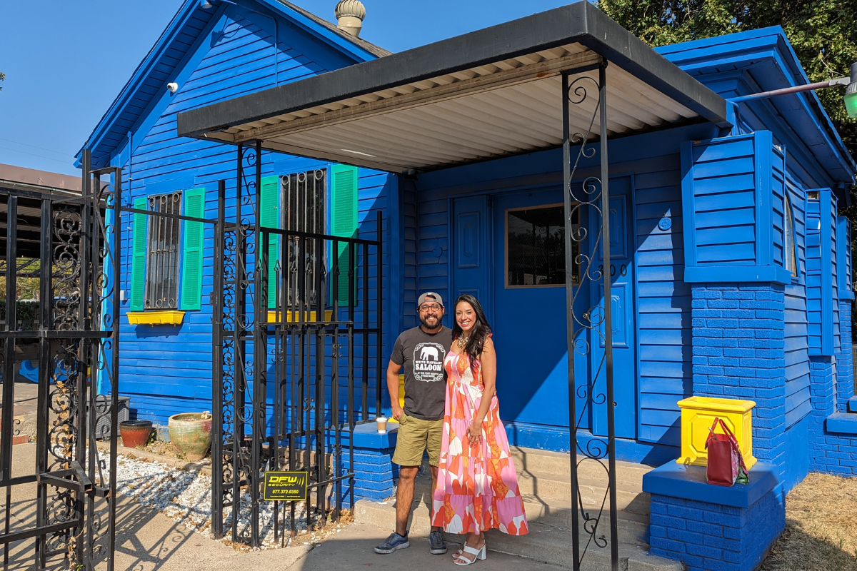 A man wearing a t-shirt and backwards cap and a woman wearing a pink dress smile in front of a blue house.