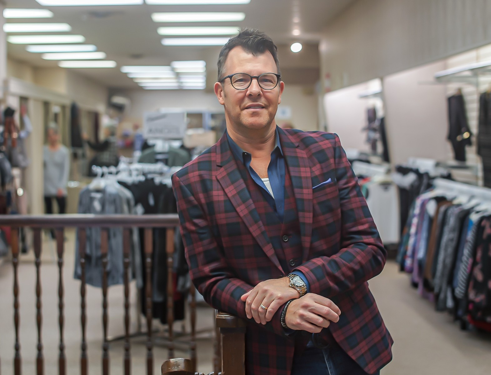 A man wearing glasses and a plaid suit jacket leans on a flight of stairs and smiles, standing in a clothing shop.