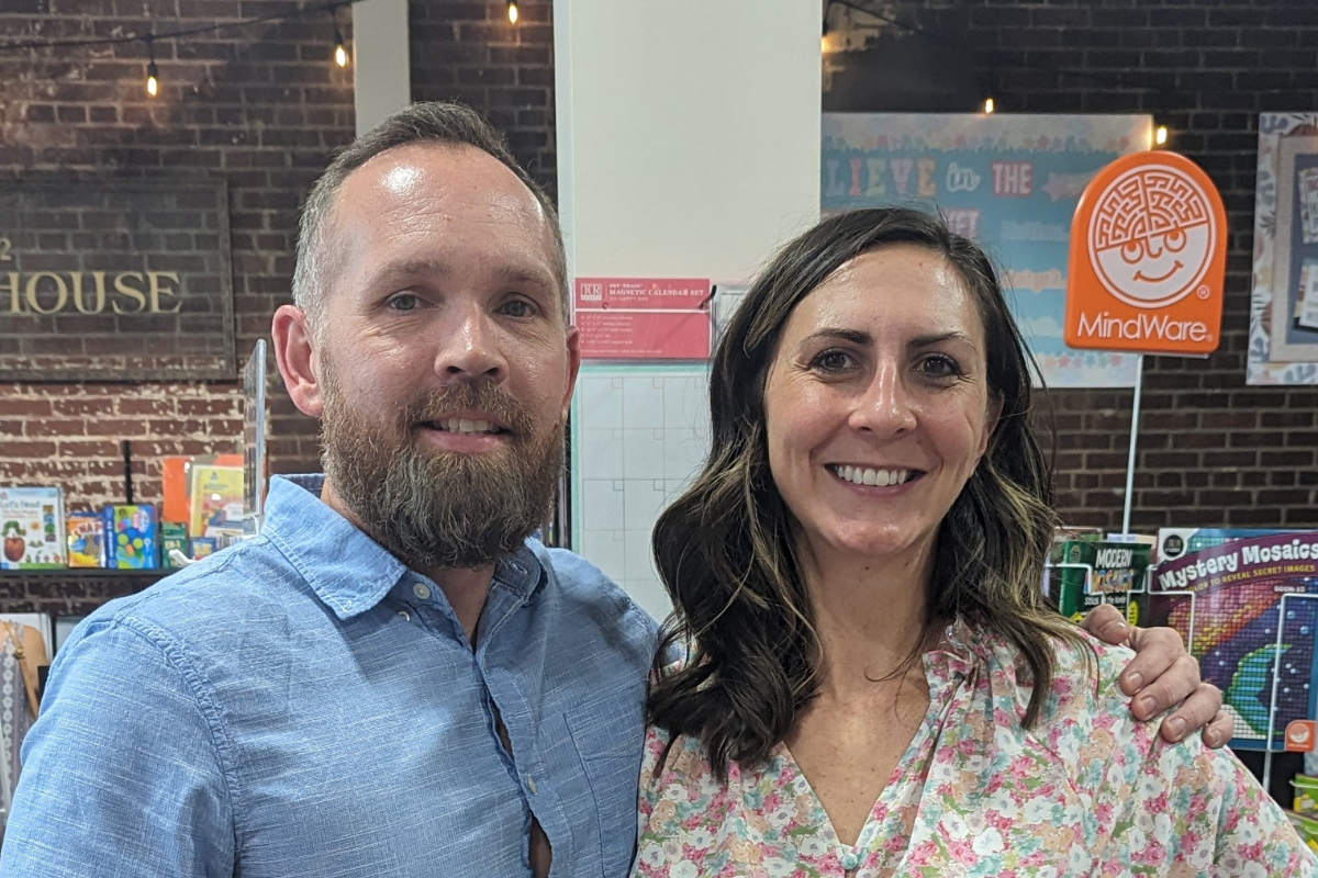 A man with a beard wearing a collared blue shirt and a woman with brown hair wearing a floral blouse smile inside a small business.