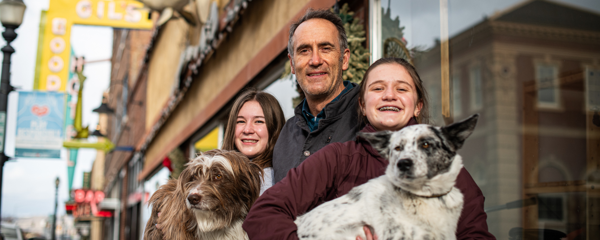 Man and two teen girls smile in front of brick building with large windows. The two girls are both holding dogs.