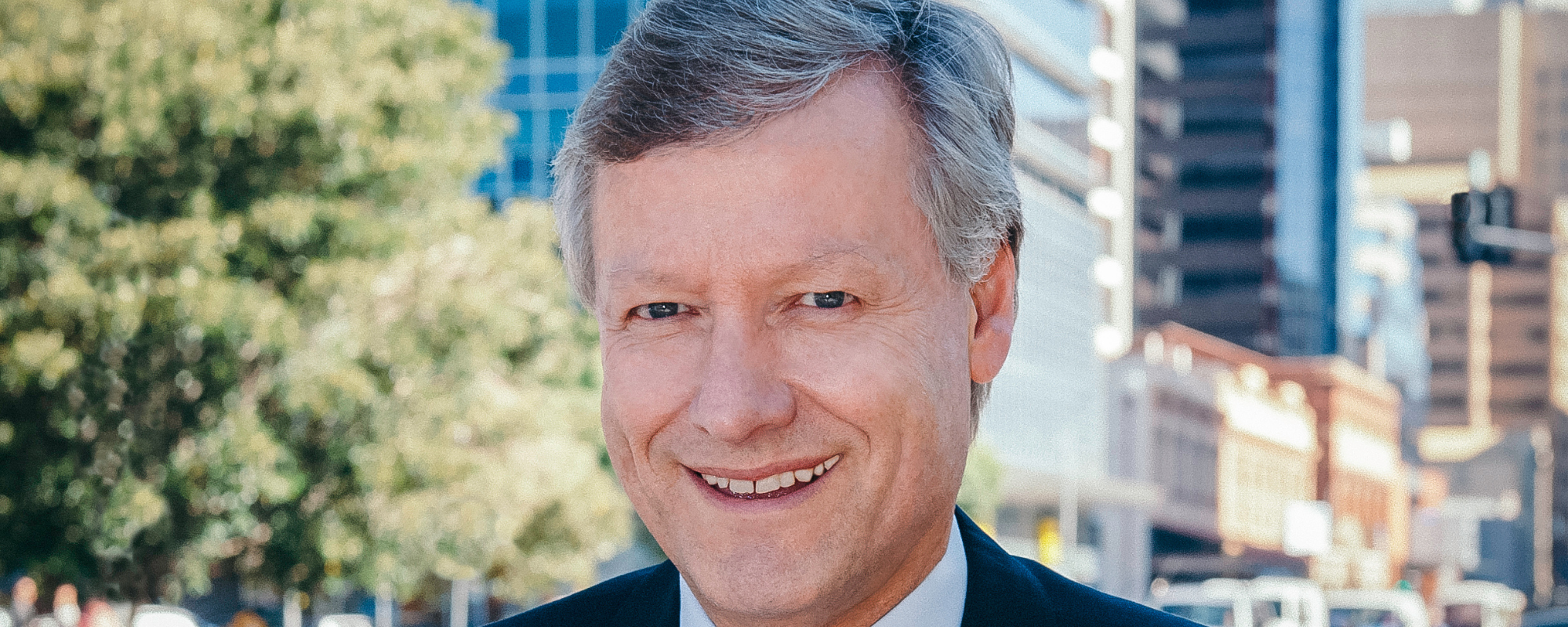 Headshot of man wearing black suit in front of city scene.