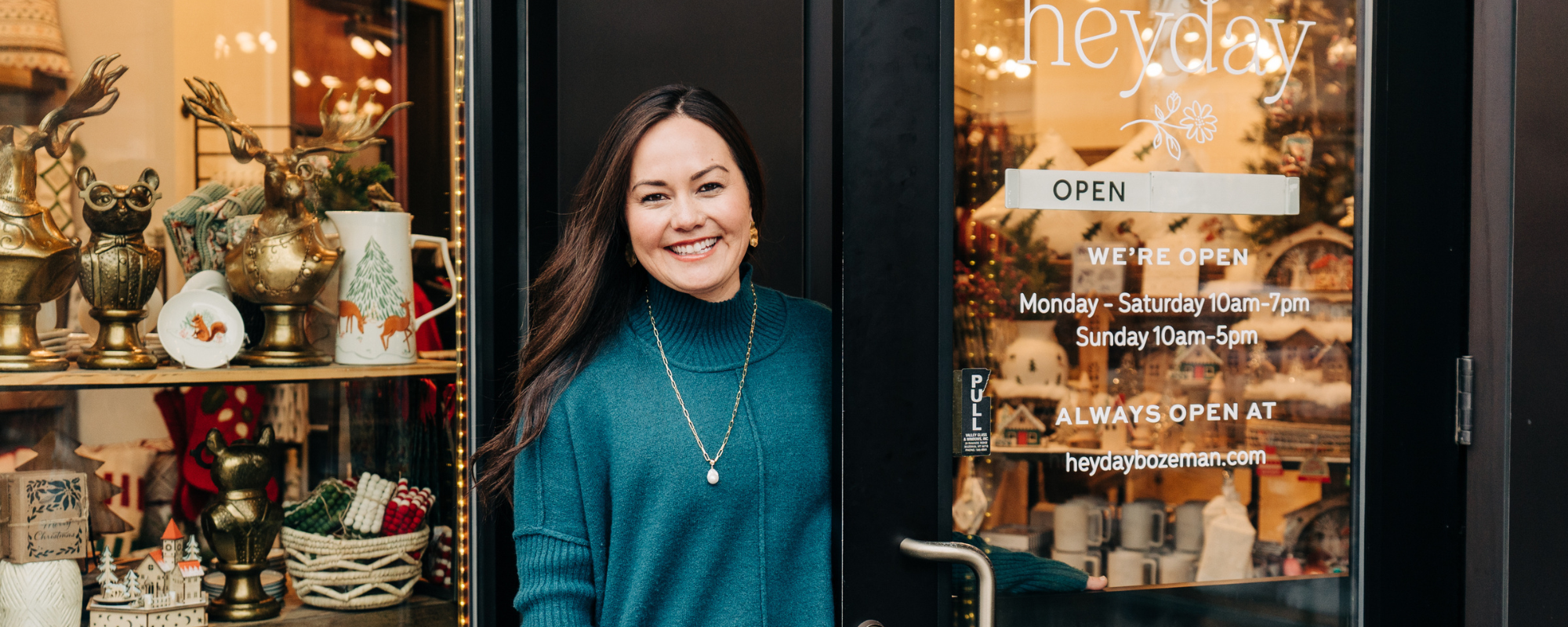 A woman wearing an emerald green sweater smiles as she opens a door to a boutique.