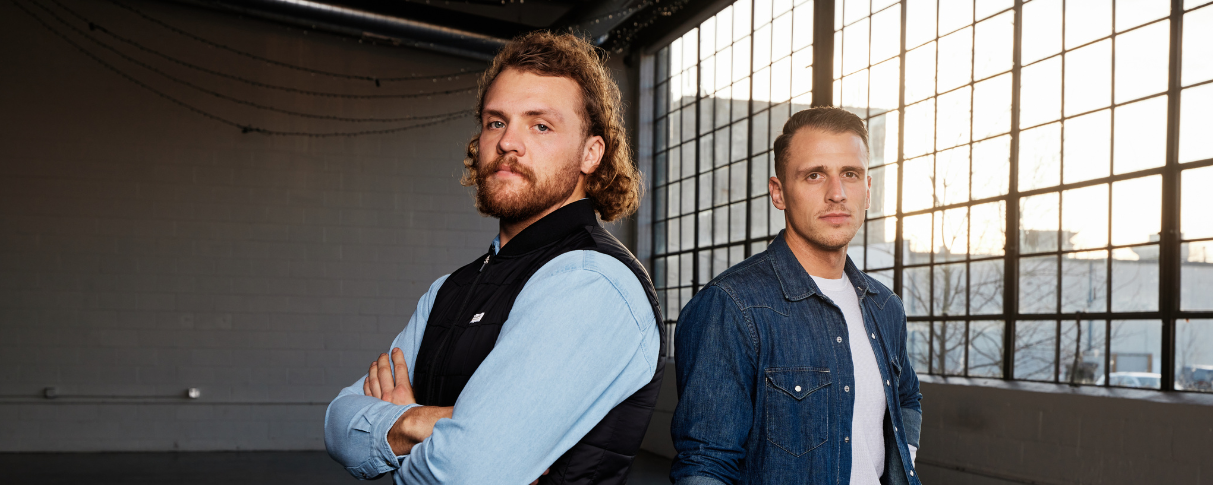 Two young men standing back to back in front of industrial window.