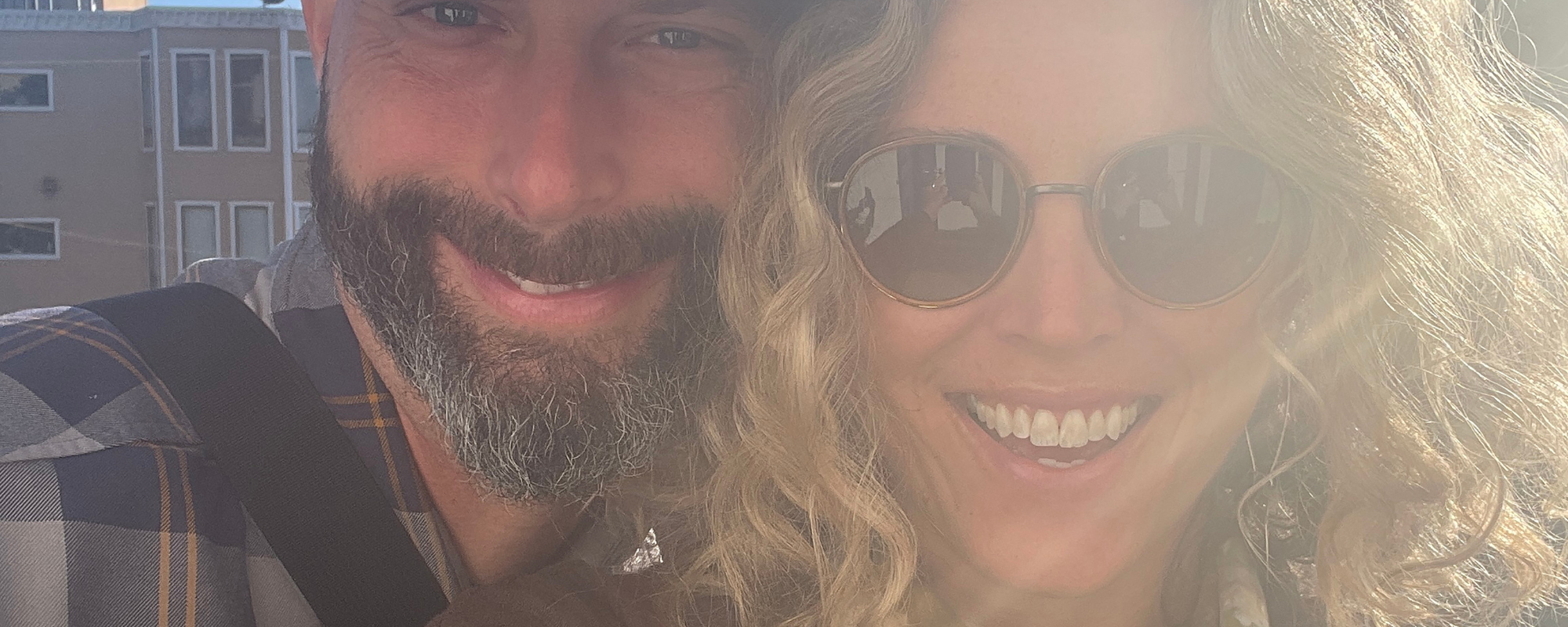 Man with beard and baseball cap smiles with woman with curly blonde hair and aviator sunglasses.