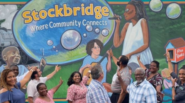 Un grupo de personas señalando un mural con burbujas, niños jugando, gente tocando música y el texto "stockbridge: where community connects" (stockbridge: donde la comunidad conecta)