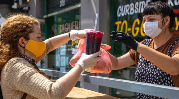 A woman wearing a face mask hands a drink and a bag of take out food to another woman wearing a face mask