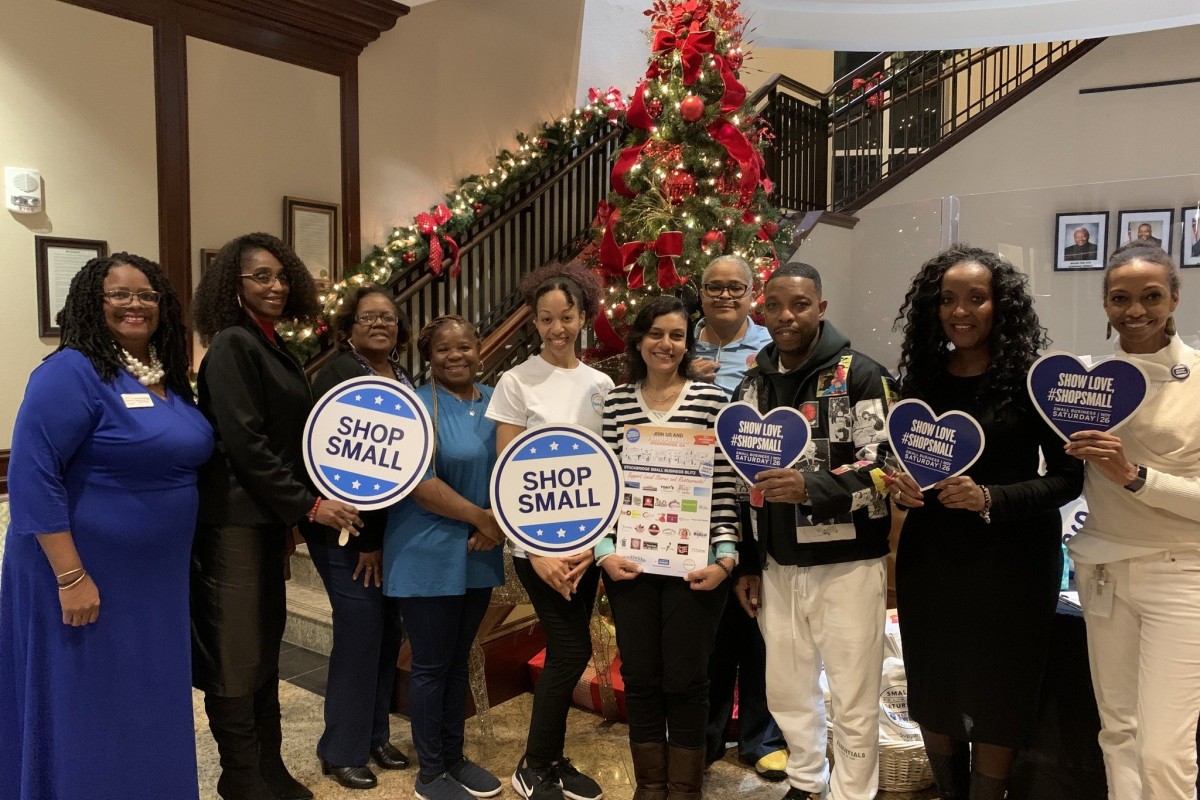 A group of people smile and hold "Shop Small" signs in front of a Christmas tree.