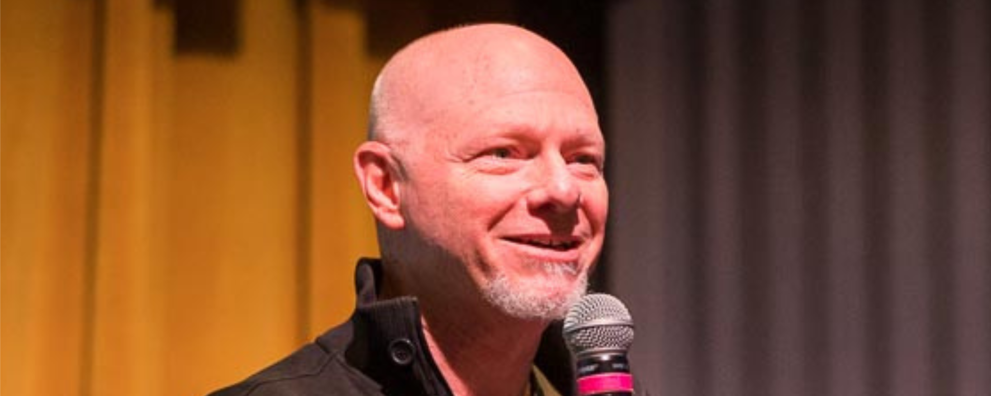 A man smiling on stage as he speaks into a microphone.
