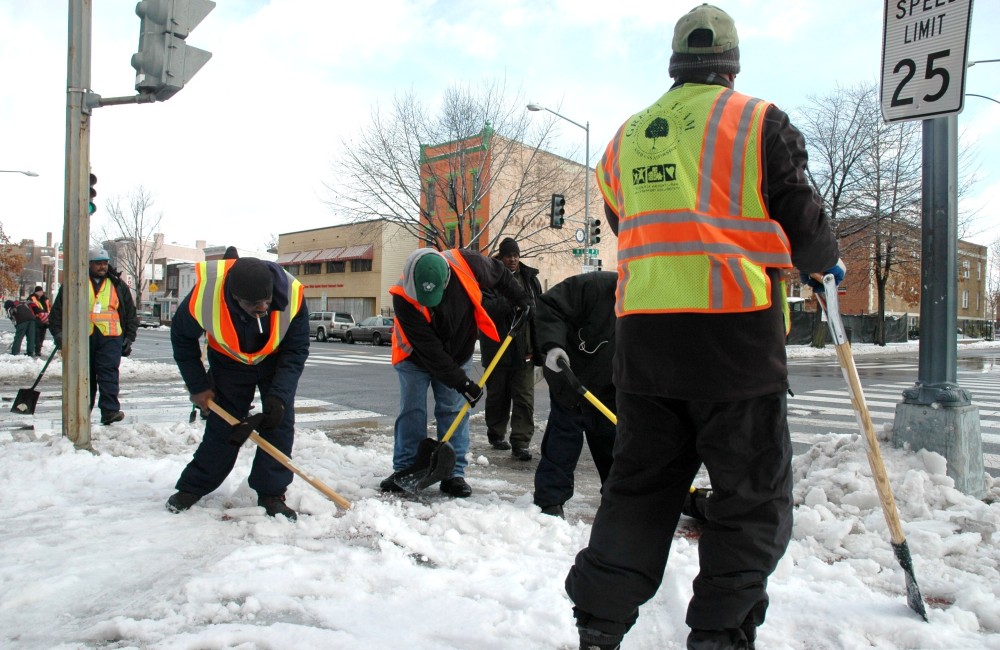 People wear hi-vis vests while shoveling show from a sidewalk.