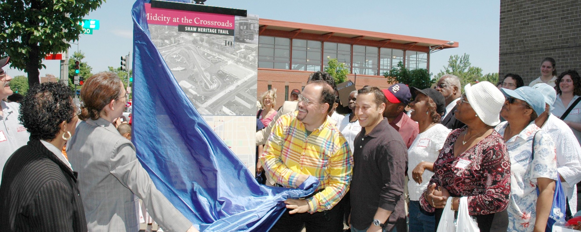 Two people remove a cloth revealing a sign for a crowd that has gathered.