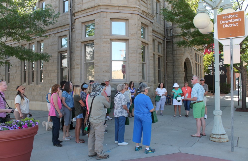 A large group of people gather around a tour guide standing in front of a large stone building.