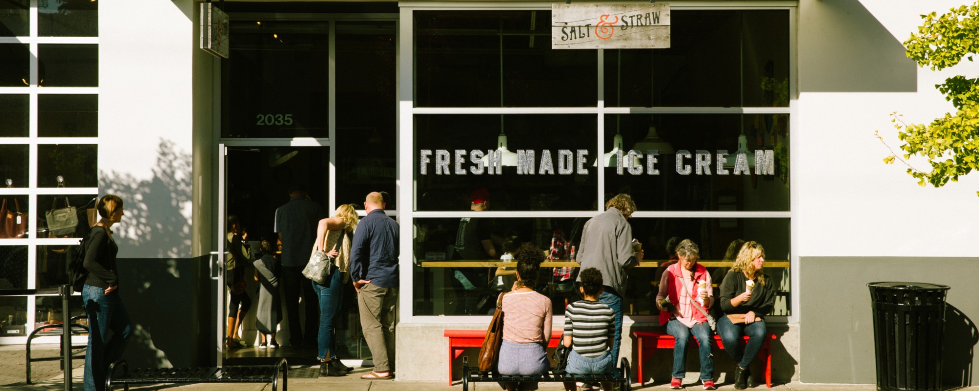 People stand in line and sit on benches outside an ice cream store.