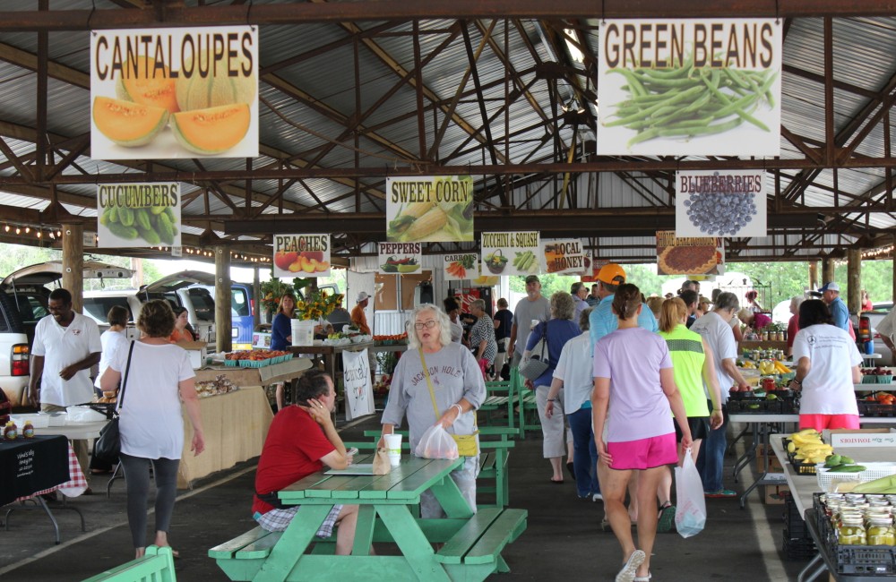People gather at brightly colored picnic tables and shop at farmers market booths set up under an outdoor pavilion.