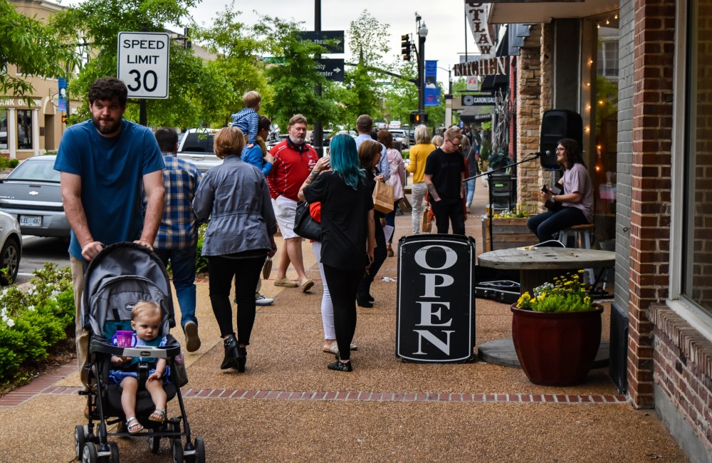 A guitarist plays and sings while people stroll along the sidewalk of a historic downtown street.