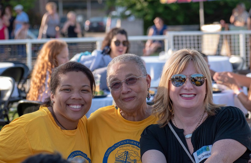 Three women smile while attending an outdoor community event.