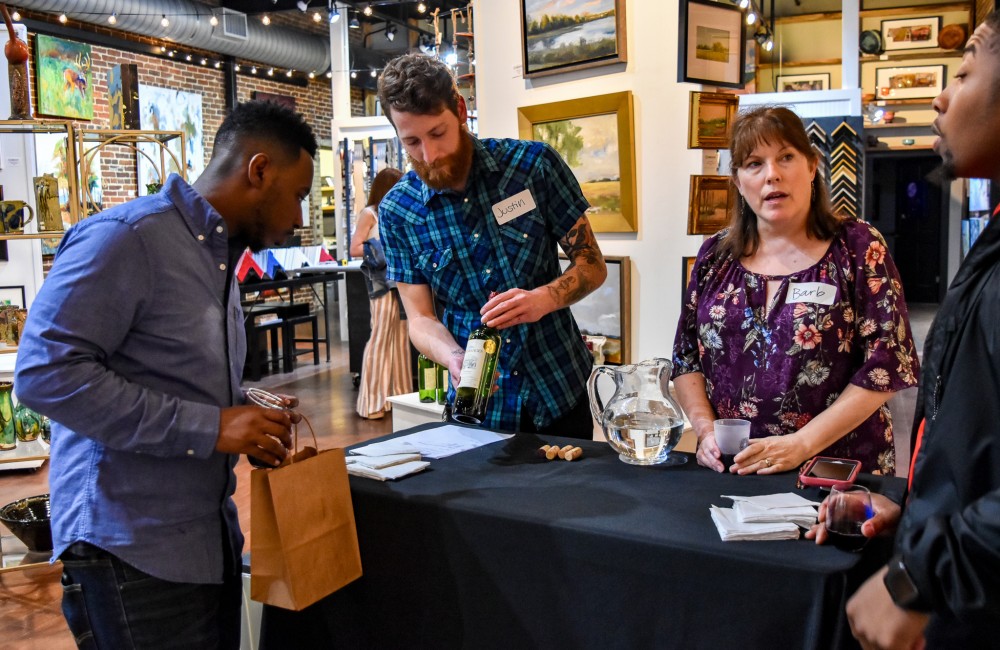 A man presents a bottle of wine to a potential customer visiting a wine tasting table set-up inside an art gallery.