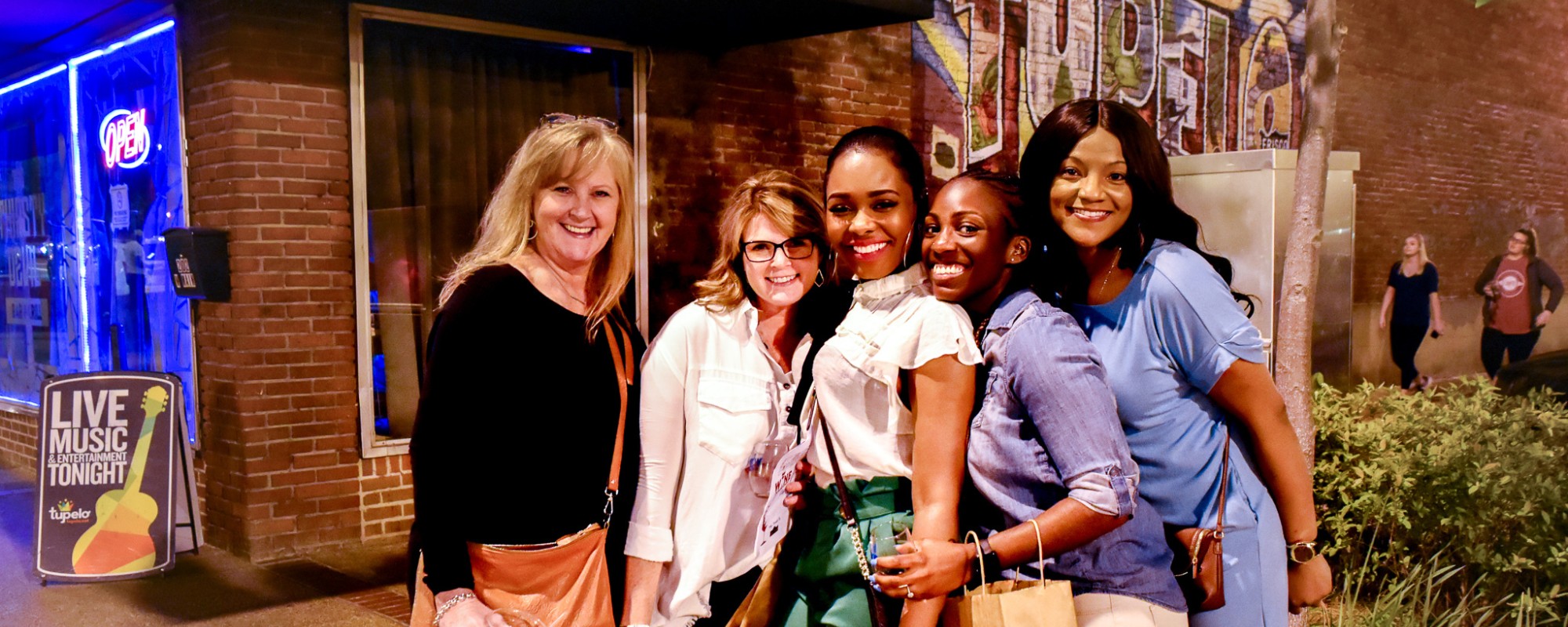 Women pose for a group photo in front of a brick building with a colorful mural.