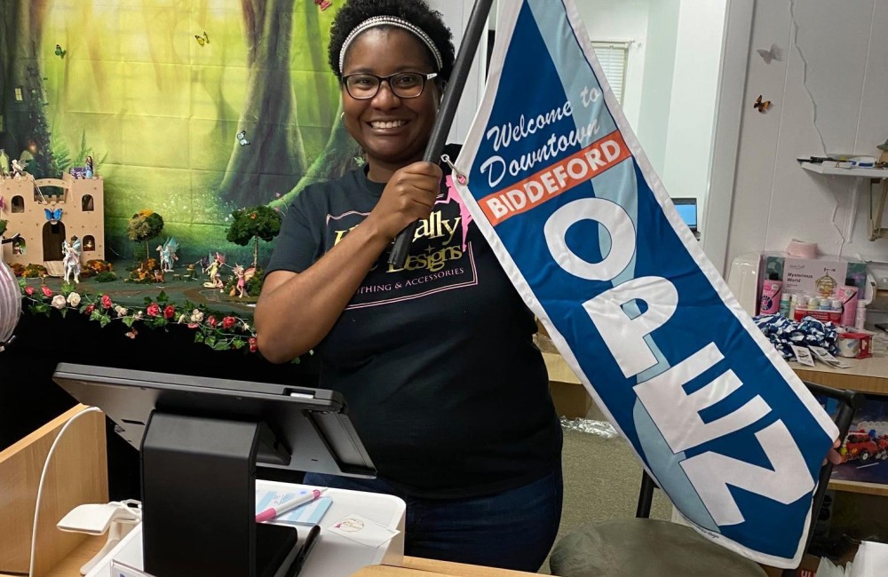 A woman stands behind her shop's check-out counter and proudly holds up her “Welcome to Downtown Biddeford” banner.