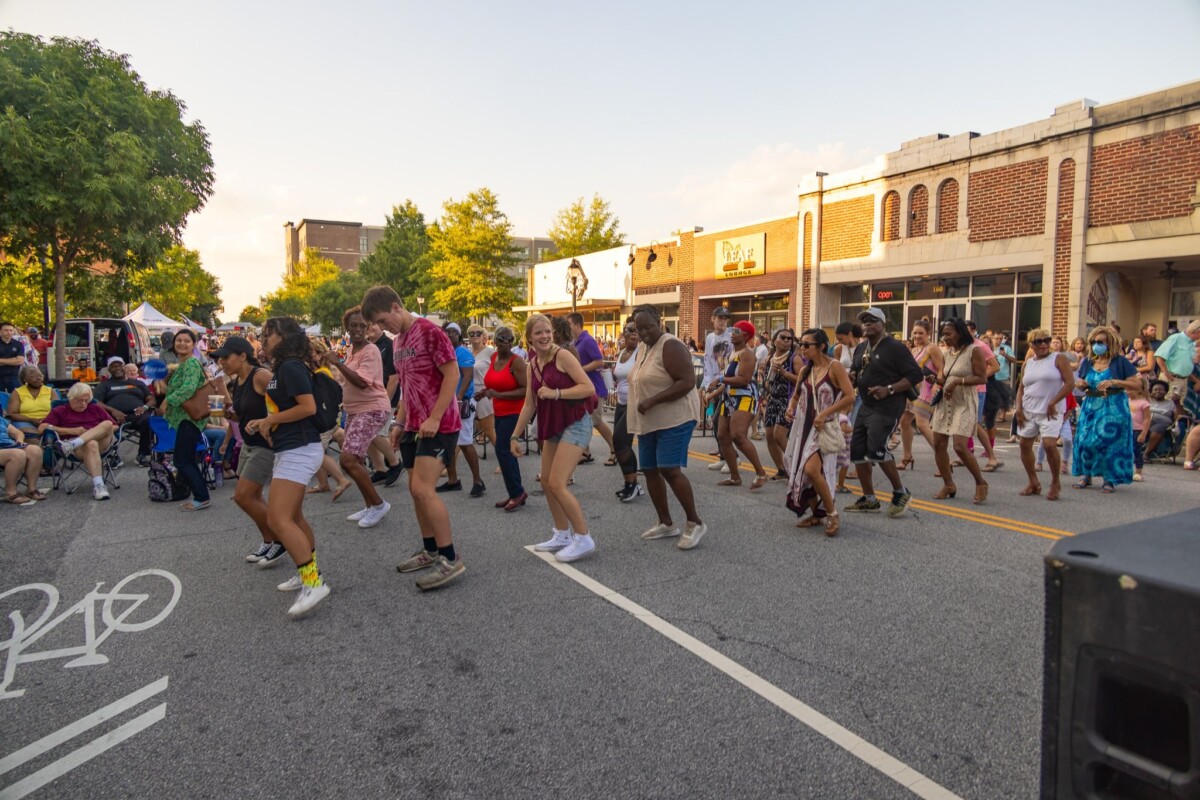 People of all ages and backgrounds line dance in the middle of a street at a summer event.
