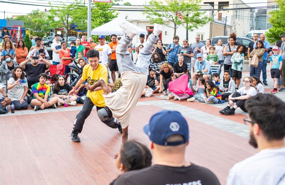 Spectators of all ages gather around an outdoor dance floor to cheer on two boys who are breakdancing.