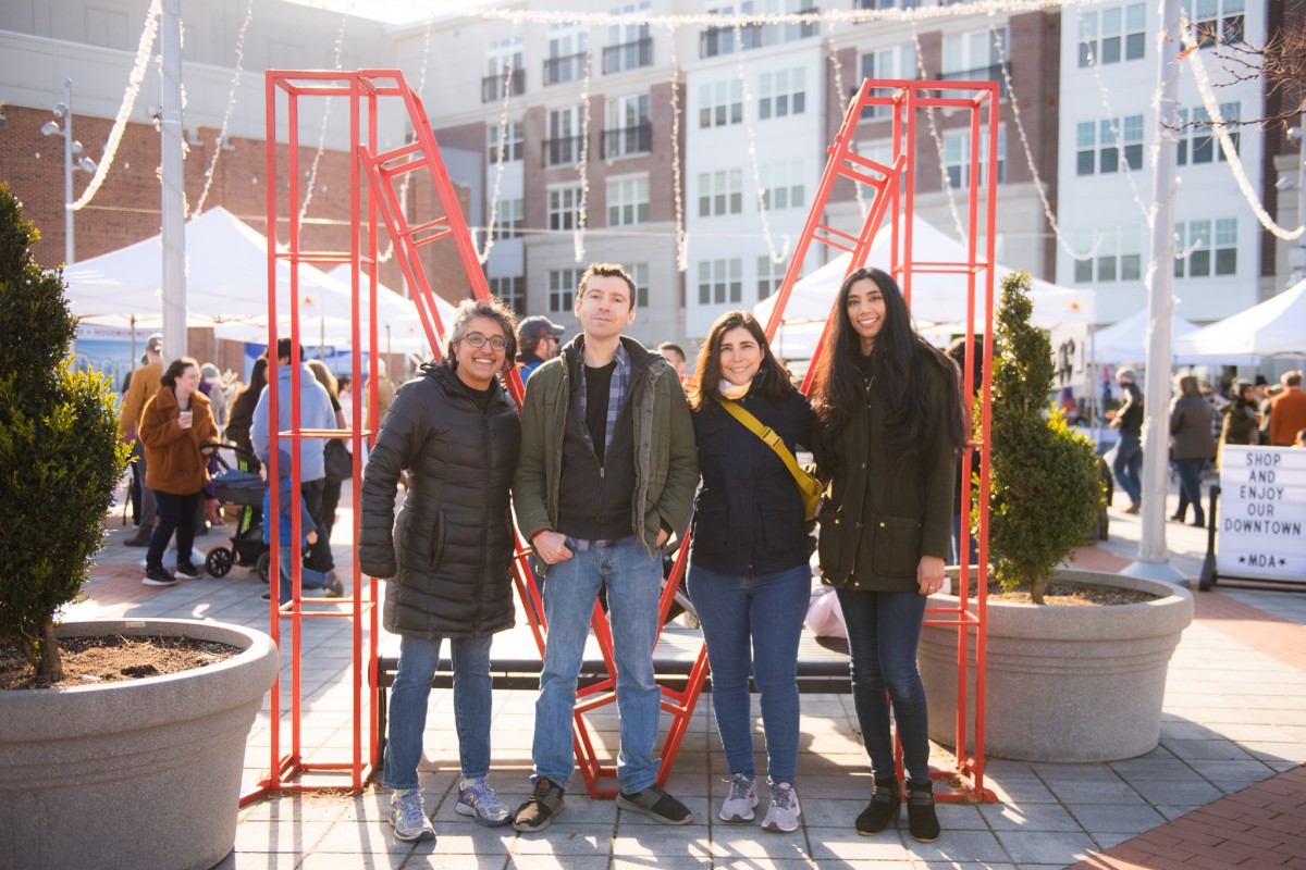 Four people stand shoulder to shoulder in front of a large red "M" structure. Behind them, a plaza is filled with visitors and white pop-up canopy tents staffed by vendors participating in a spring bazaar event.