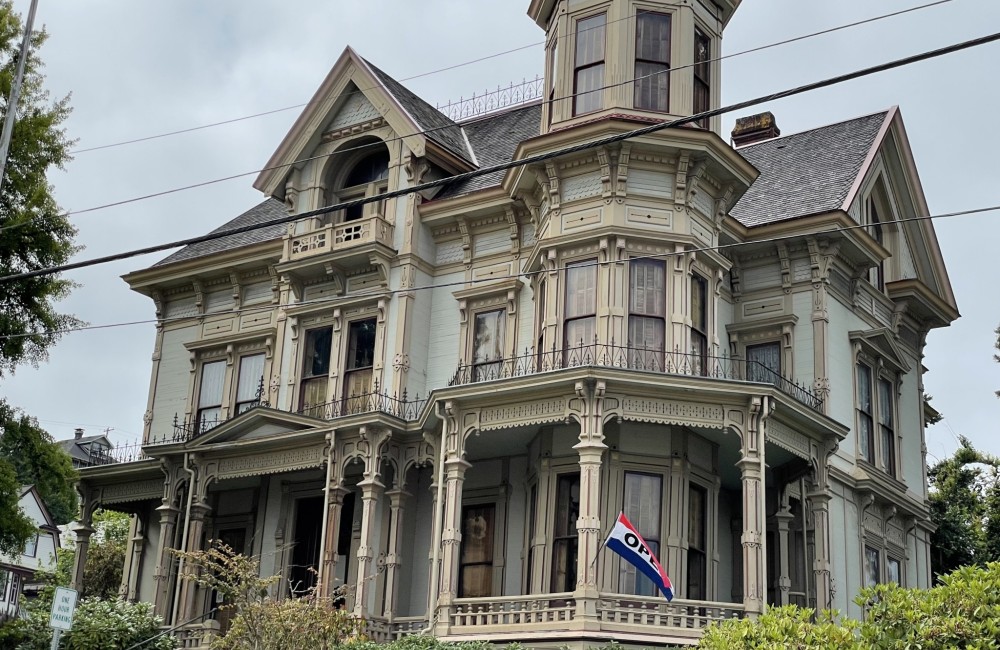 A large, ornate Queen Anne-style house with intricate architectural details featuring a prominent tower with multiple windows, decorative woodwork, and a wrap-around porch supported by elaborate columns. The exterior is painted in muted tones of cream and beige with dark trim.