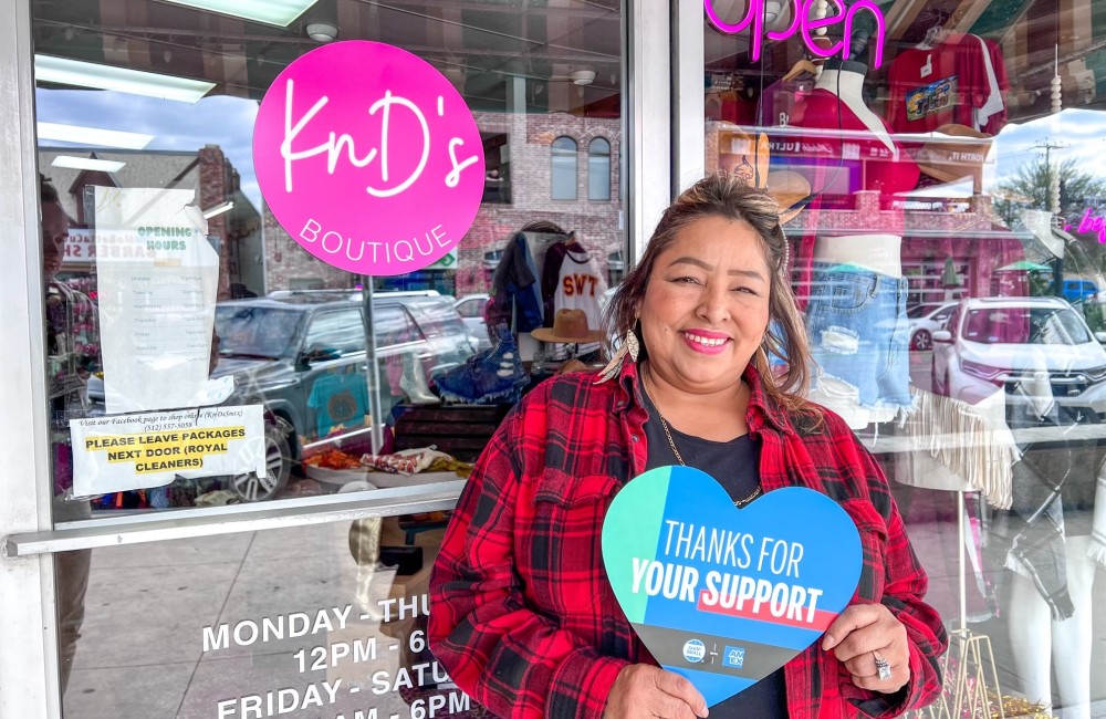 A person is holding a sign shaped like a heart that says, "Thanks for your support" while smiling in front of a business that has a decal that says, "KnD's Boutique".