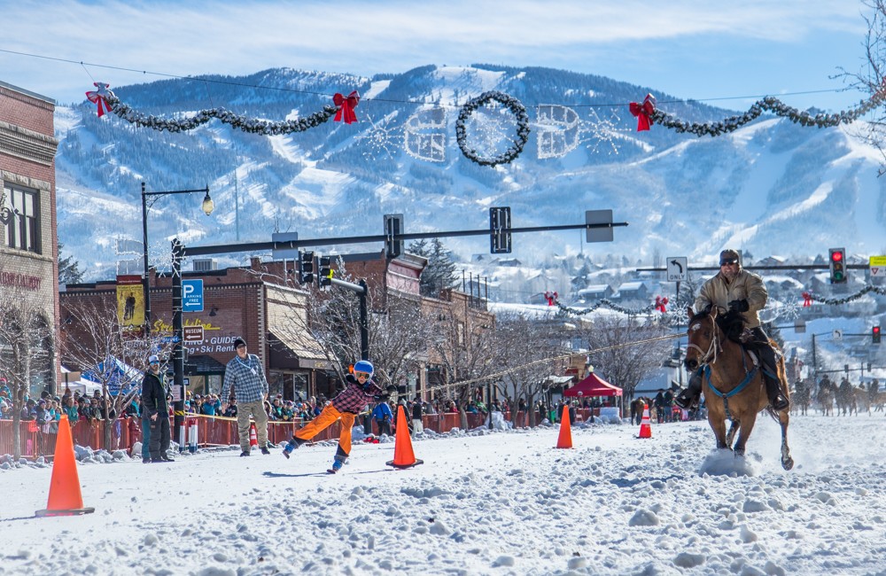 A child is holding onto a rope attached to a man riding a horse down a snow covered street.