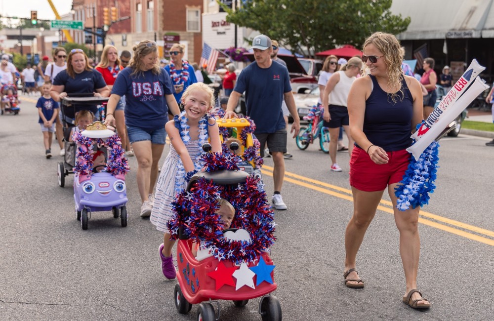 A child is smiling pushing another child in a wagon on the street during a parade.