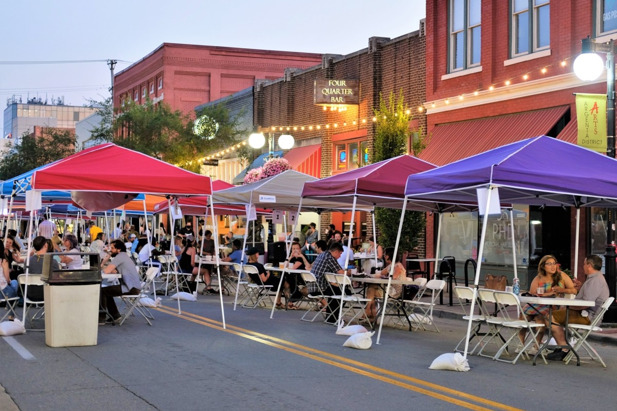 People gather around tables set-up underneath brightly colored pop-up canopy tents that have been arranged in a single line down the middle of a downtown street.