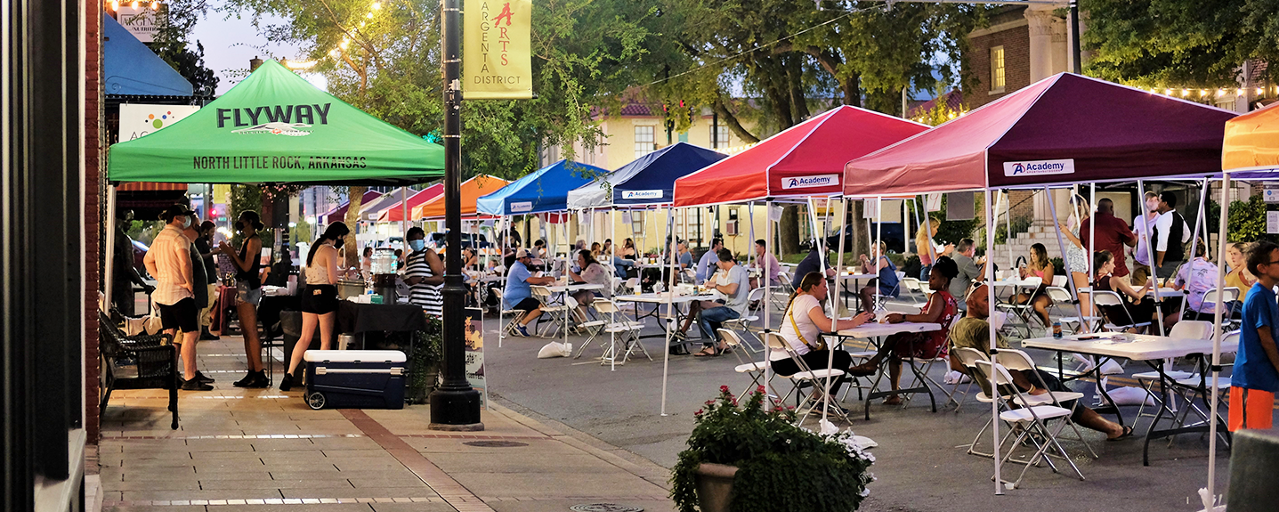 People gather around tables set-up underneath brightly colored pop-up canopy tents that have been arranged in a single line down the middle of a downtown street.