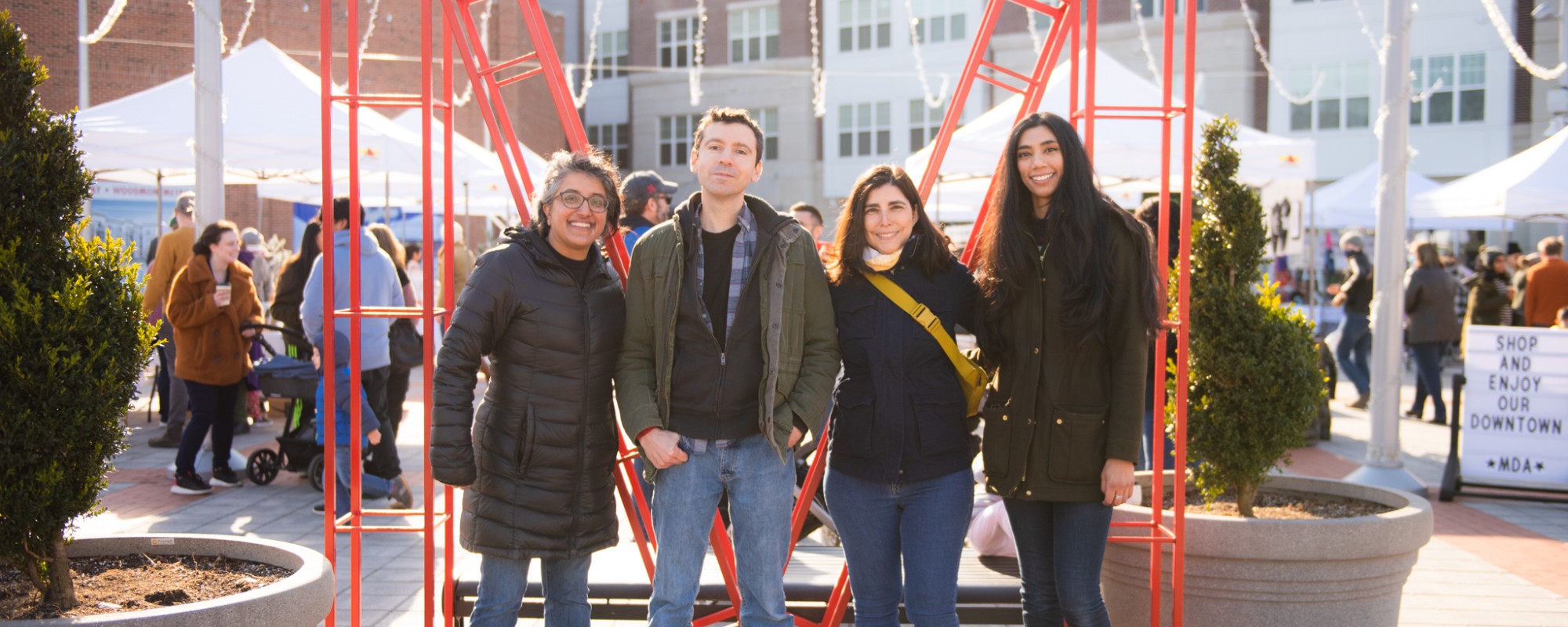 Four people stand shoulder to shoulder in front of a large red "M" structure. Behind them, a plaza is filled with visitors and white pop-up canopy tents staffed by vendors participating in a spring bazaar event.
