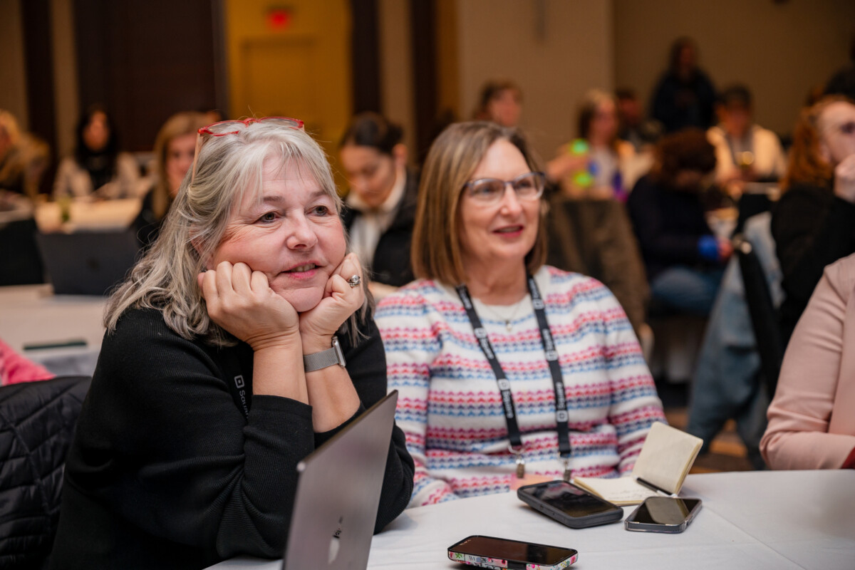 Two women are sitting at a table and listening intently to a speaker.