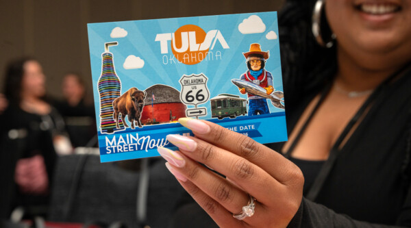 A woman holding up a "save the date" postcard for the 2026 Main Street Now Conference.