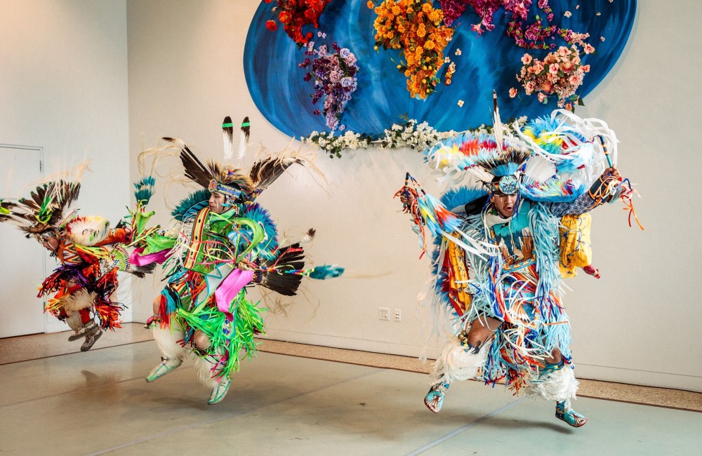Three Native American dancers dressed in colorful regalia dance in an indoor space. On the wall behind them, a large installation depicting the globe with continents made from flowers is visible.