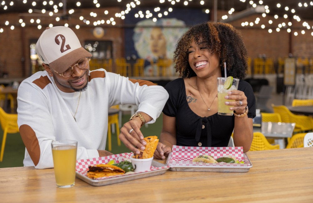 A man and woman enjoy a meal at an indoor marketplace with ample seating and twinkle lights hanging from the ceiling.